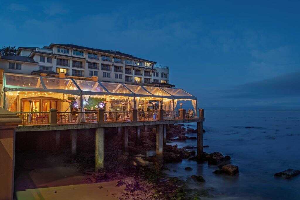 Brightly lit restaurant overlooking the ocean at dusk, located on a rocky cliff's edge in California.