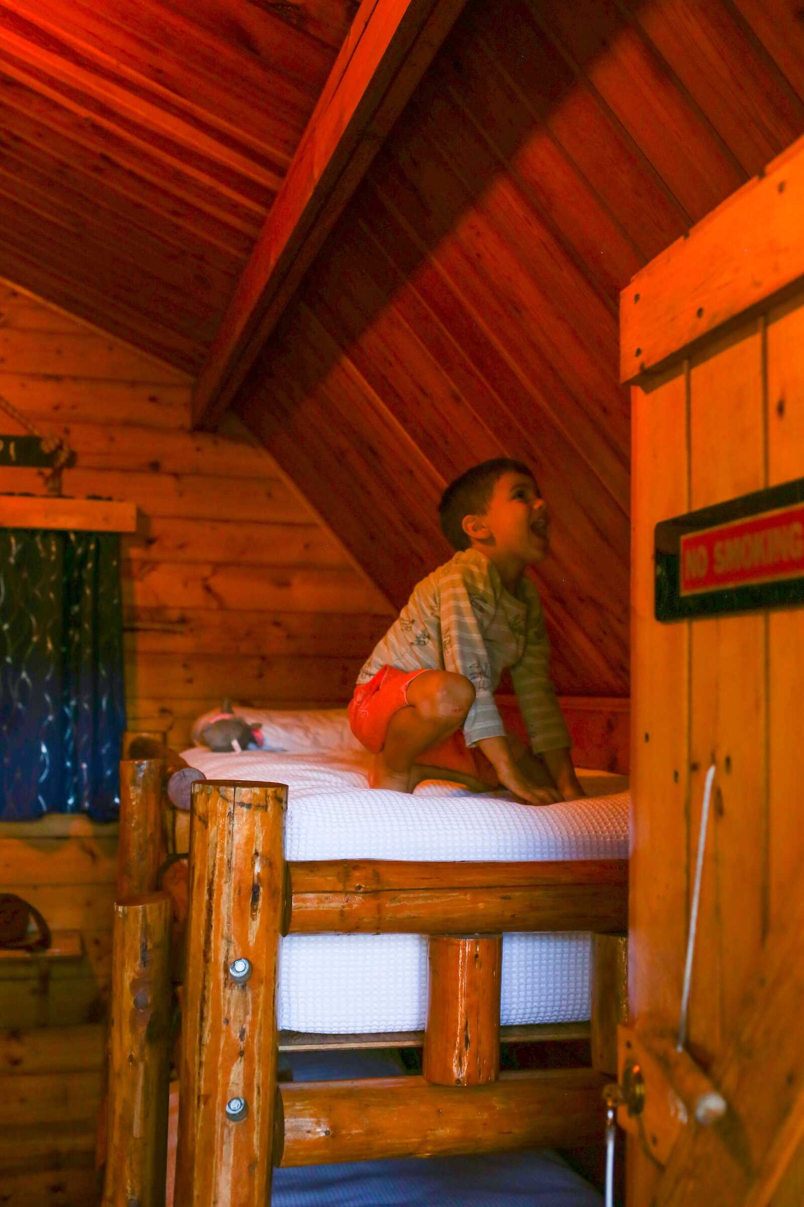 Cozy wooden cabin bedroom with a child playing on the bed, rustic interior, warm lighting, and adventure vibe.