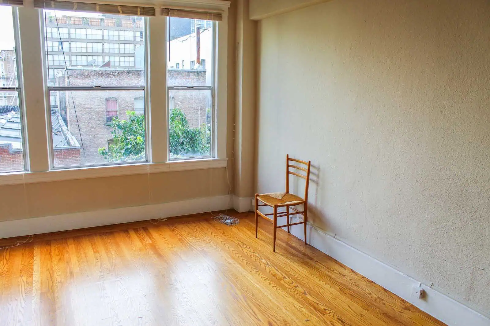 Empty room with large window overlooking city buildings, wooden floor, and single wooden chair.