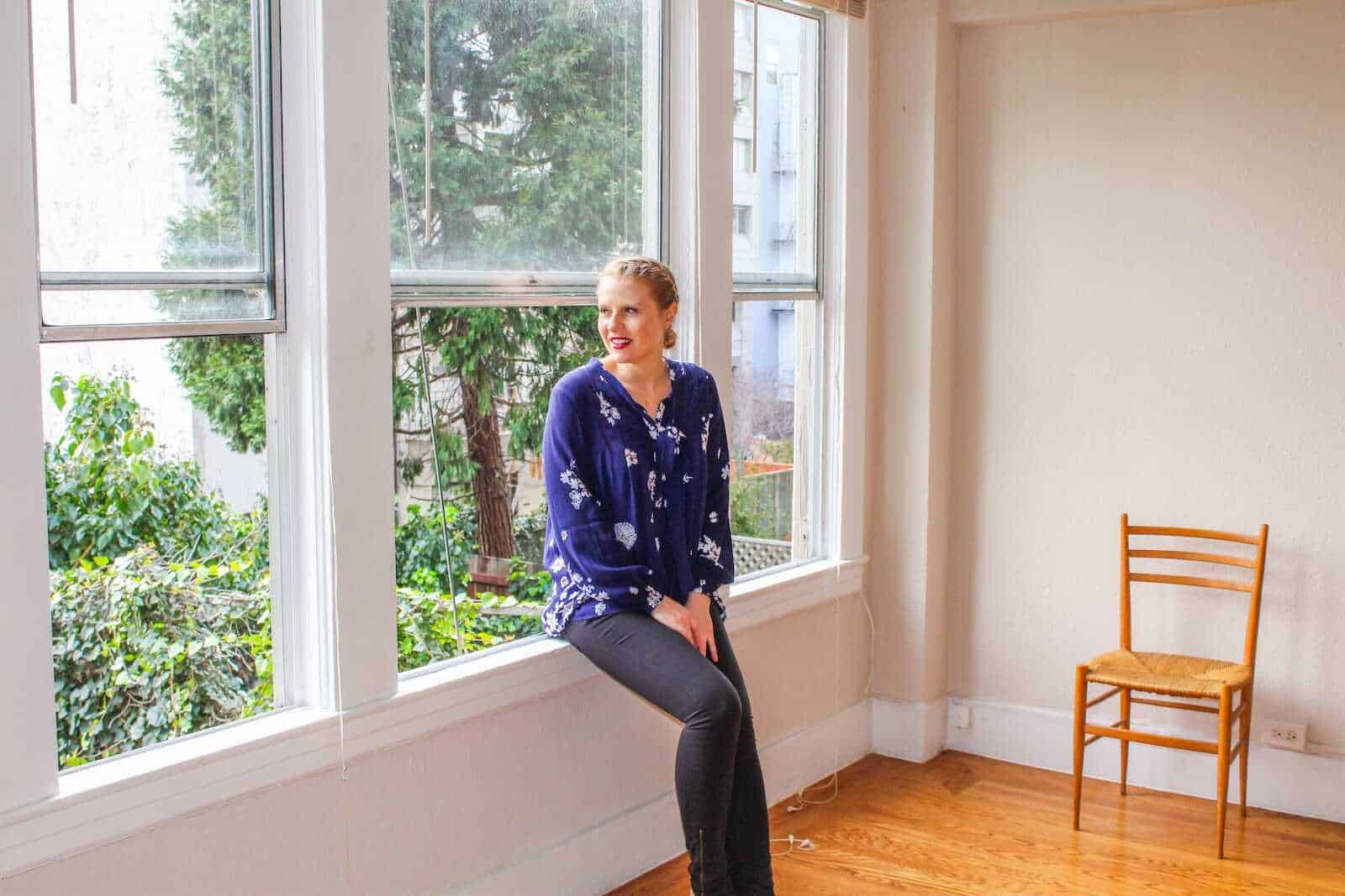 Serene woman relaxing by large window in cozy California apartment, surrounded by greenery.